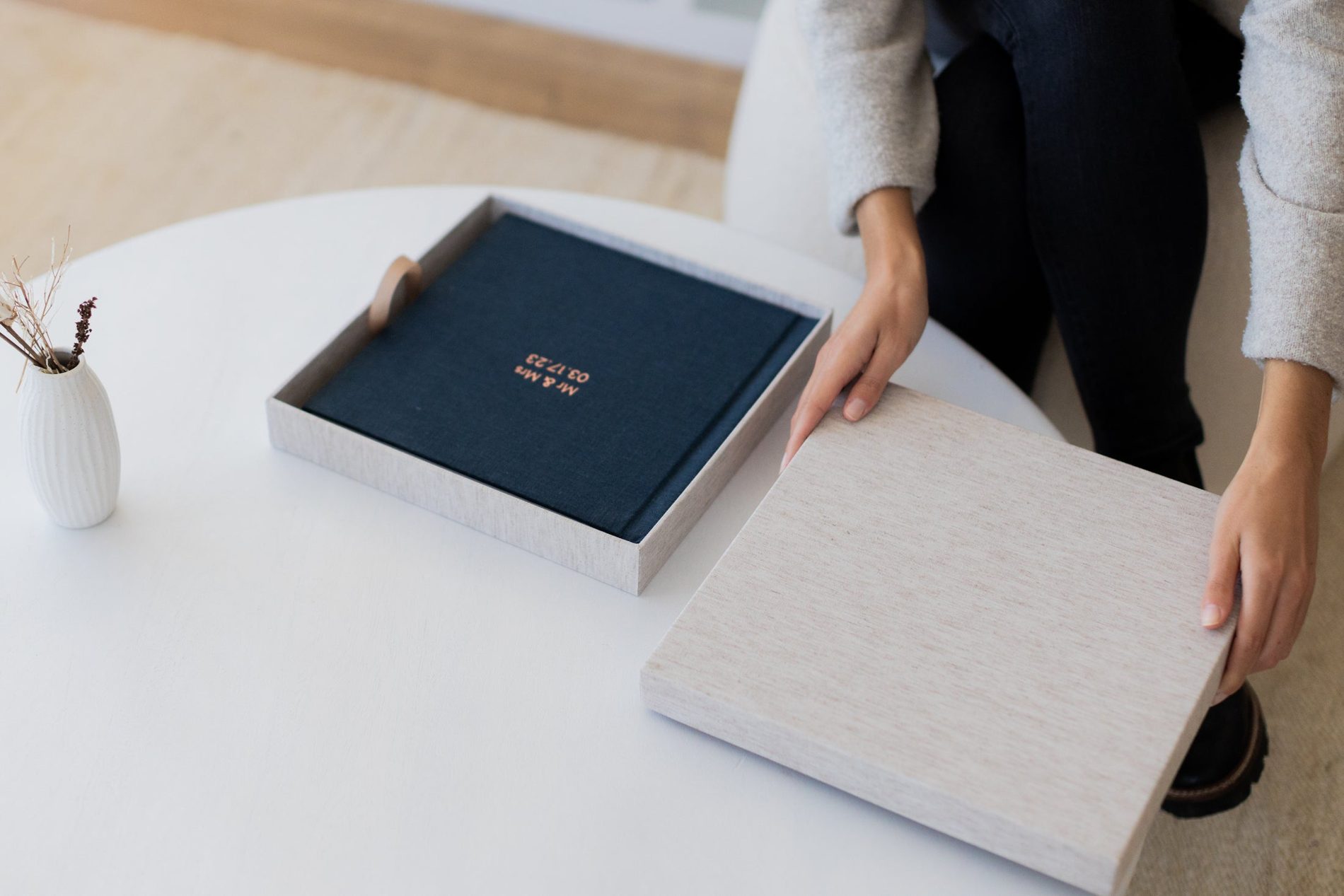 A client lifting the lid off a linen presentation box on a coffee table, a navy linen album with foil lettering inside