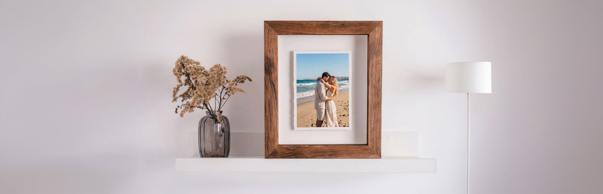 A dark wood frame on a white shelf, holding a hand-torn print of a couple embracing on a beach