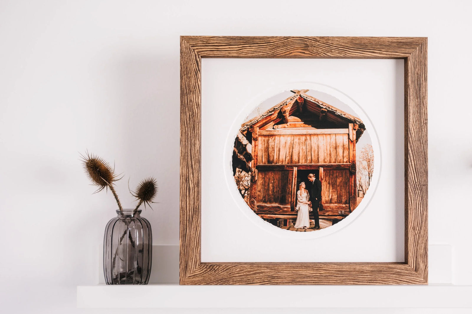 A brown wood frame with a white mat, holding a round hand-torn print of a couple in front of a wooden barn
