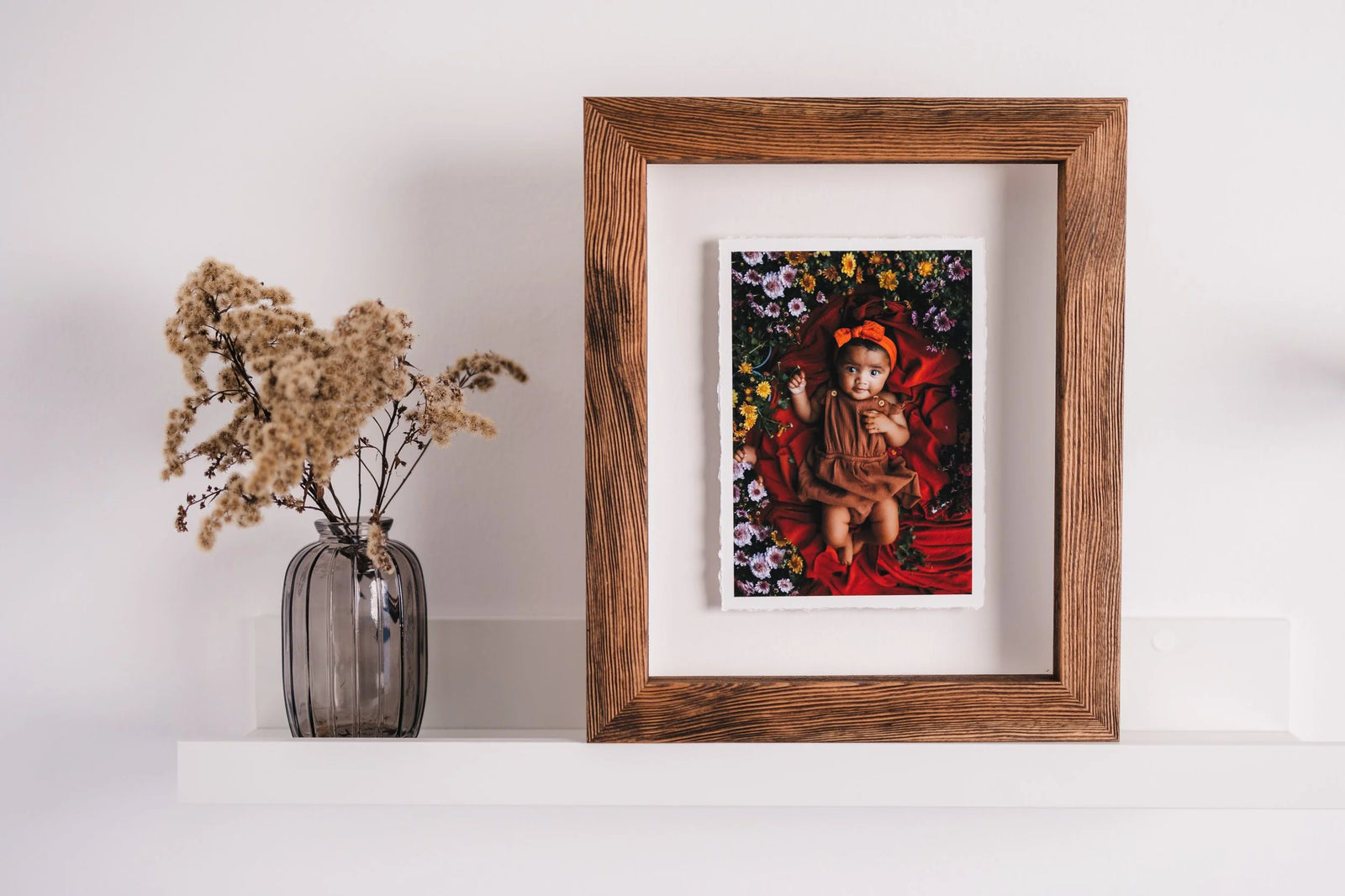 A wood frame holding a hand-torn print of a baby lying among flowers, raised above the white mount